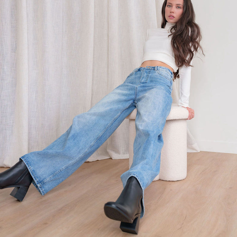 Model wearing OKAKIE Adelle high-rise wide-leg denim jeans in a vintage wash with a white turtleneck and black boots.