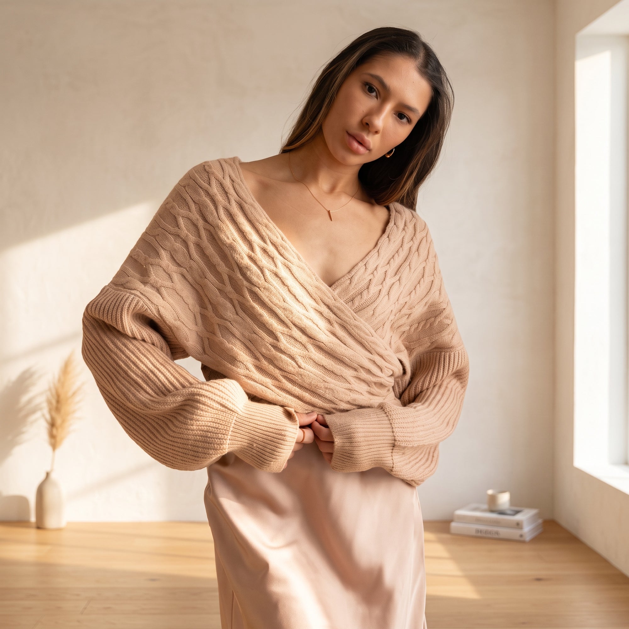 Model wearing an OKAKIE beige cable knit sweater with a criss cross V-neckline and wide dolman sleeves in a bright room