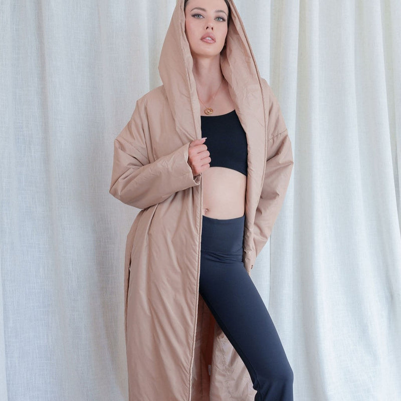 Model wearing an OKAKIE beige longline pillow puffer coat with an oversized hood and relaxed dropped shoulders.