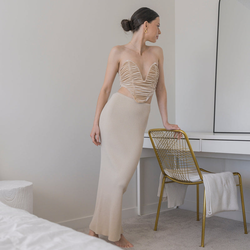 OKAKIE beige ribbed knit maxi skirt worn by a woman in a minimalist bedroom setting with a gold chair.