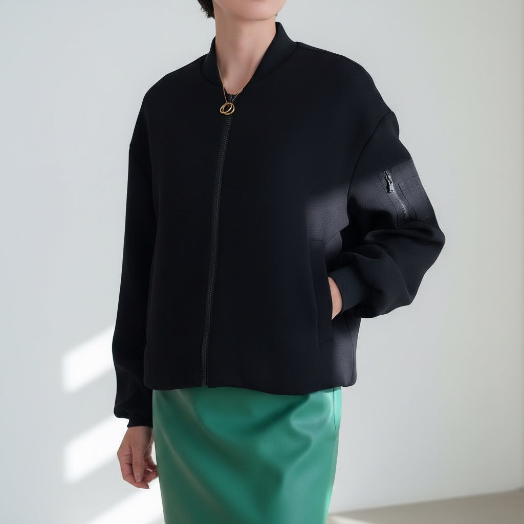 OKAKIE black bomber jacket with a front zipper, ribbed collar, and utility sleeve pocket worn over a green skirt.