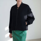 OKAKIE black bomber jacket with a front zipper, ribbed collar, and utility sleeve pocket worn over a green skirt.