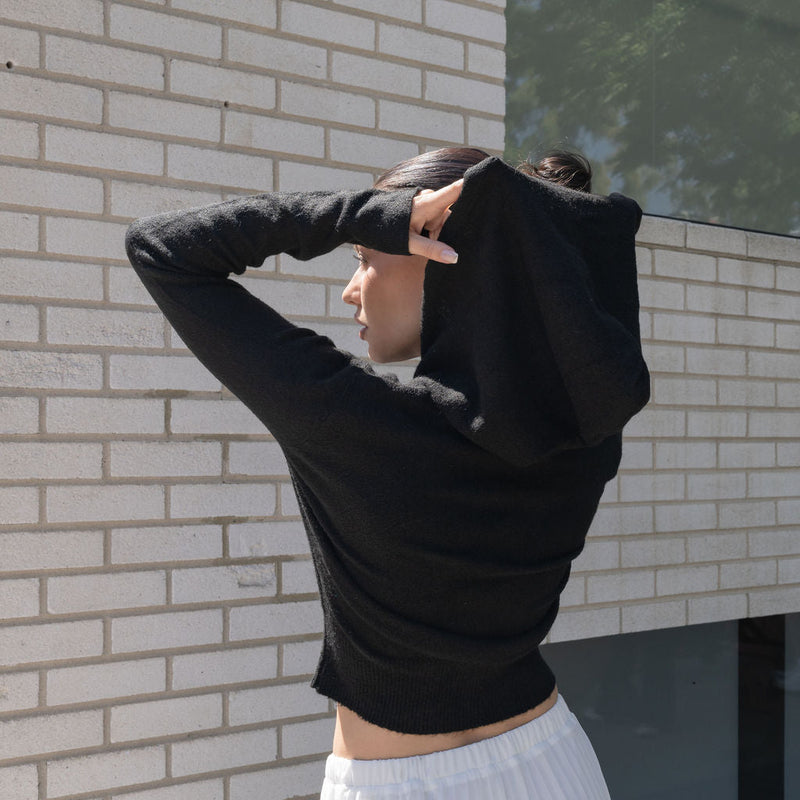 OKAKIE black cotton knit zip hoodie with hood up, worn by a woman against a white brick wall