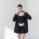 Model wearing an OKAKIE black cotton poplin boyfriend over-shirt over a matching corset top and mini skirt