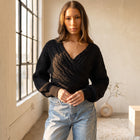 OKAKIE black cable knit sweater with a criss-cross V-neckline and dolman sleeves worn by a model in a bright studio.