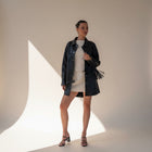 Model wearing an OKAKIE black vegan leather shacket with fringe sleeves over a cream mini dress in a studio setting.