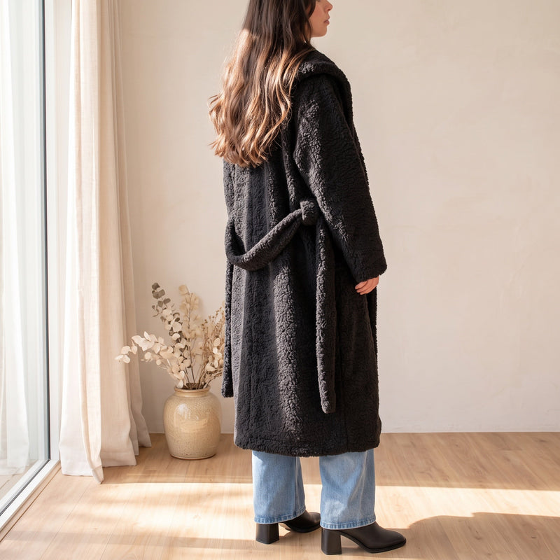 OKAKIE black plush teddy maxi robe coat with a belted waist, worn over blue jeans in a bright, minimalist room.