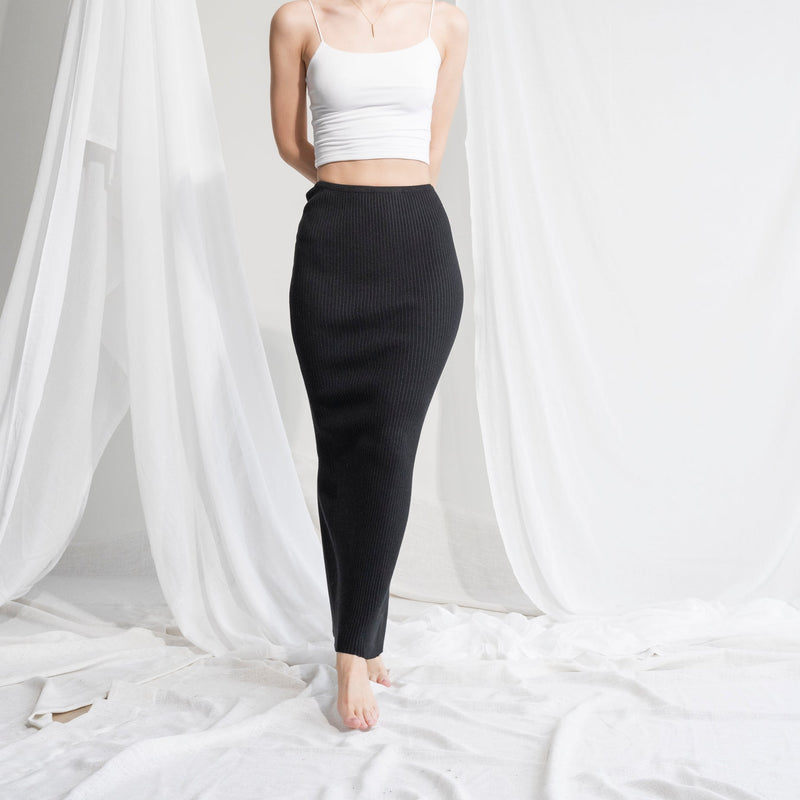 OKAKIE black ribbed knit maxi skirt worn with a white camisole against a draped white fabric background.