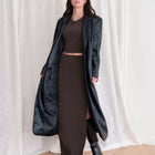 OKAKIE longline black vegan leather trench coat worn over a dark brown matching set with knee-high boots.