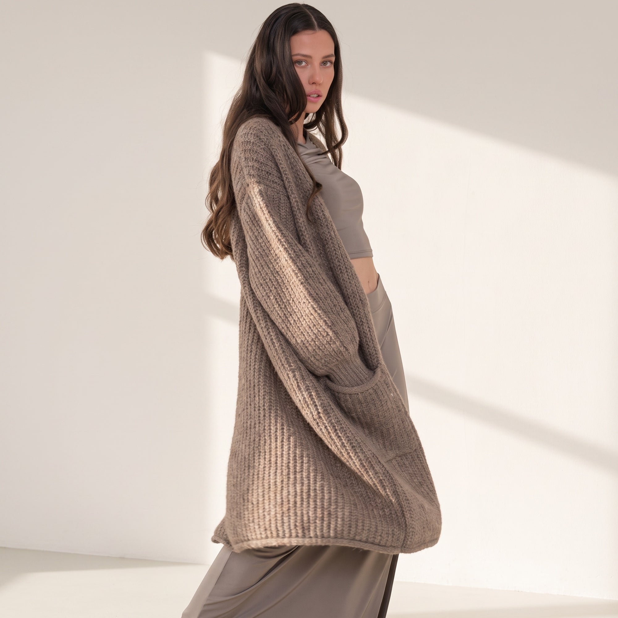 Model wearing an OKAKIE heavy warm brown purl knit cardigan with long sleeves and front pockets in a sunlit studio.