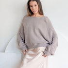 Model wearing OKAKIE beige oversized reversible sweater with dropped shoulders and a wide neckline over a silk skirt.
