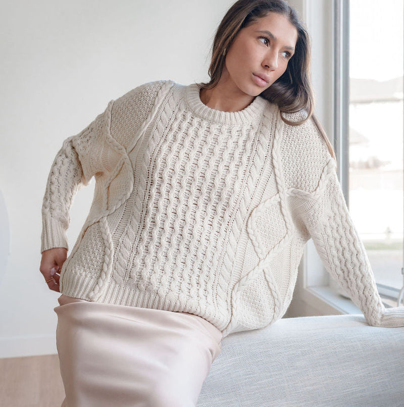 OKAKIE cream oversized cable knit sweater with mixed textures and crew neckline worn by a woman sitting on a sofa
