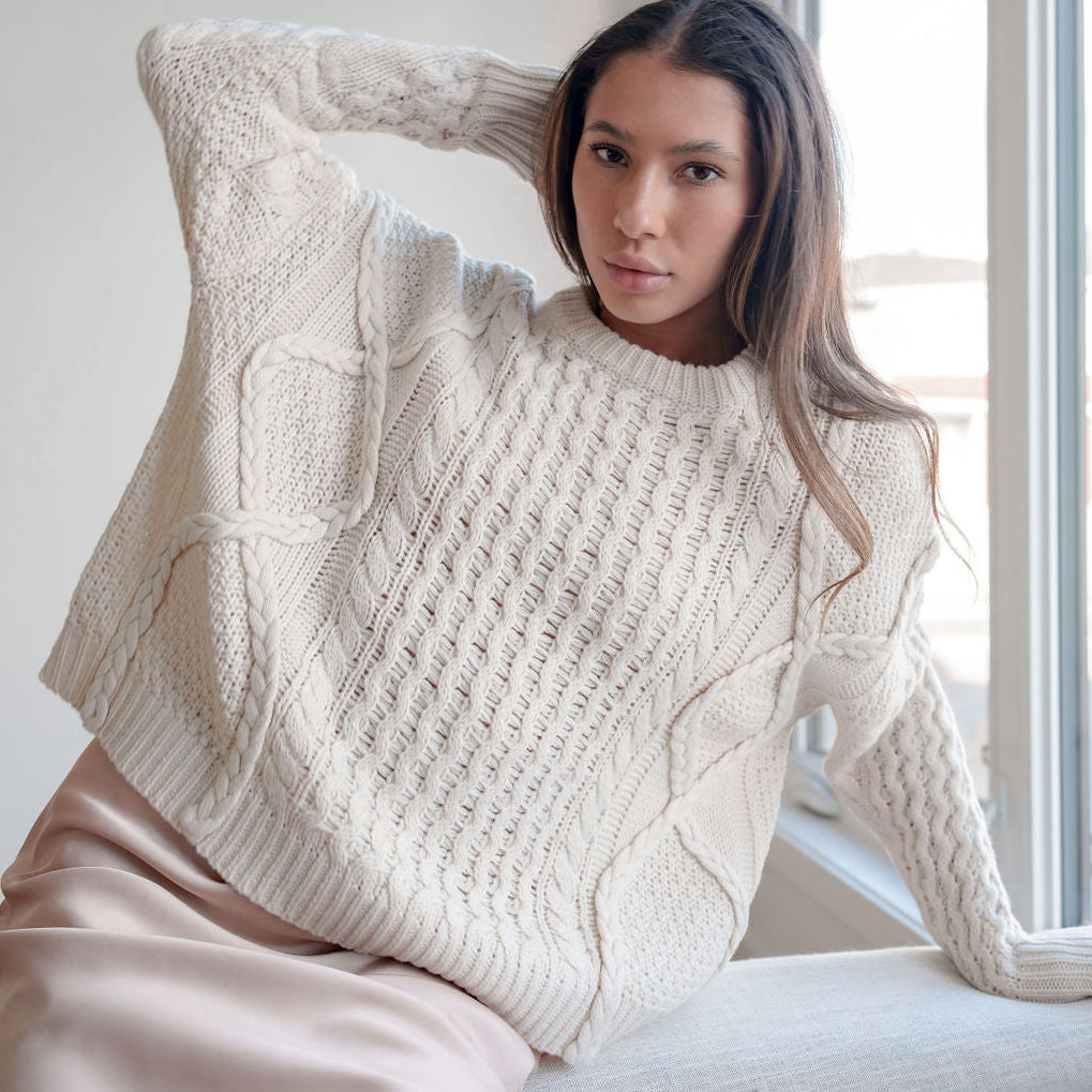 OKAKIE cream oversized cable knit sweater with mixed textures and crew neck worn by a woman sitting by a window