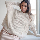 OKAKIE cream cable knit sweater with oversized fit, crew neck, and dropped shoulders worn by a woman indoors.
