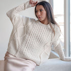 Model wearing an OKAKIE cream heavyweight cotton cable-knit sweater with a relaxed oversized fit and crew neckline.