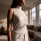 Model wearing OKAKIE cream sleeveless high neck rib knit top with curved detailing in a sunlit living room