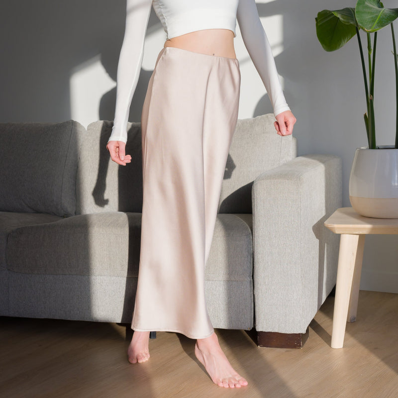 OKAKIE Essential maxi skirt in champagne matte satin worn with a white long-sleeve top in a sunlit living room.