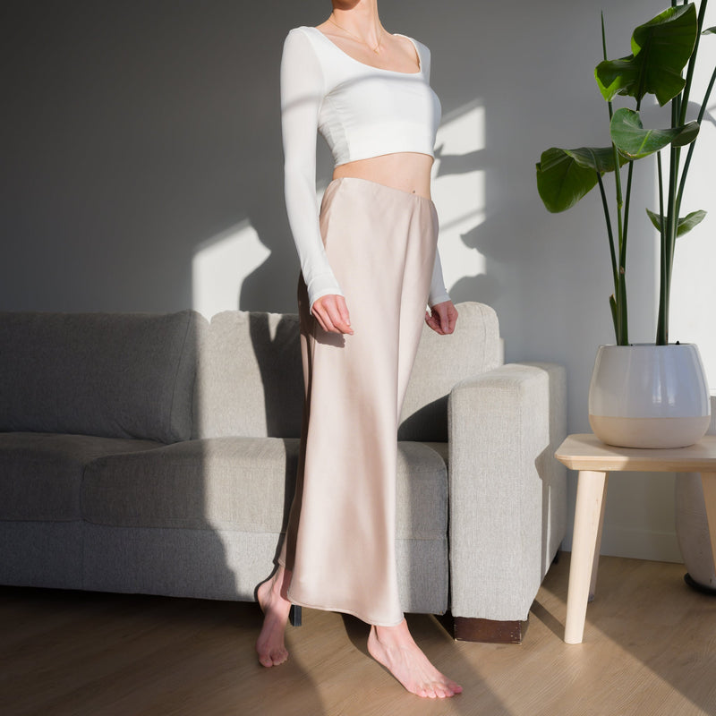 OKAKIE matte satin maxi skirt in champagne, worn with a white long-sleeve crop top in a sunlit living room.