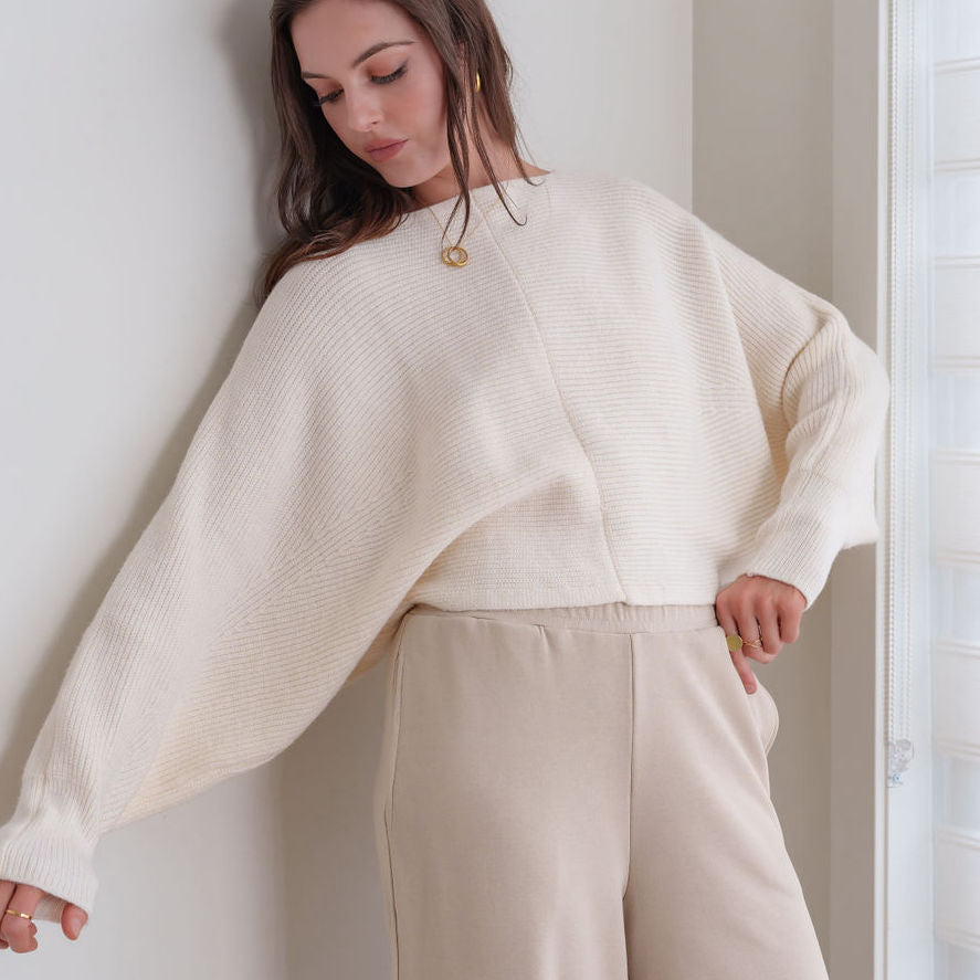 OKAKIE ivory knit sweater with a relaxed fit, wide sleeves, and ribbed cuffs worn by a woman against a neutral wall.