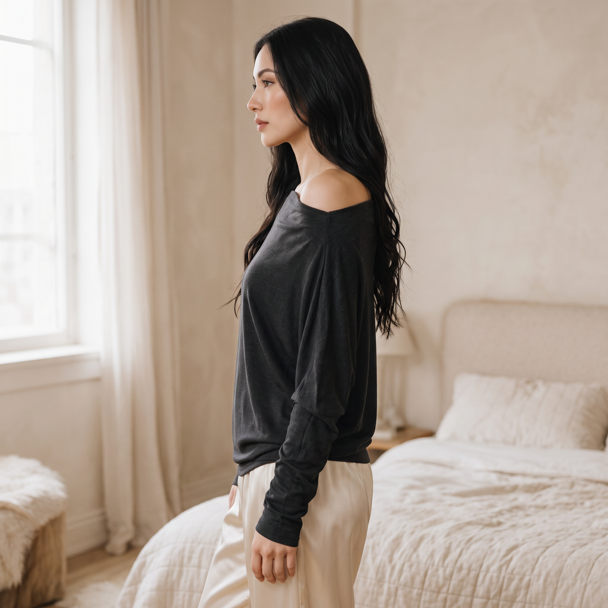 OKAKIE Maëlle draped charcoal hacci knit top with a wide boat neckline worn off-shoulder in a soft bedroom setting.