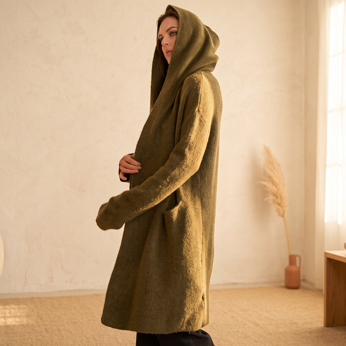 Model wearing an OKAKIE olive heavyweight hooded cardigan with an open front, longline silhouette, and patch pockets.