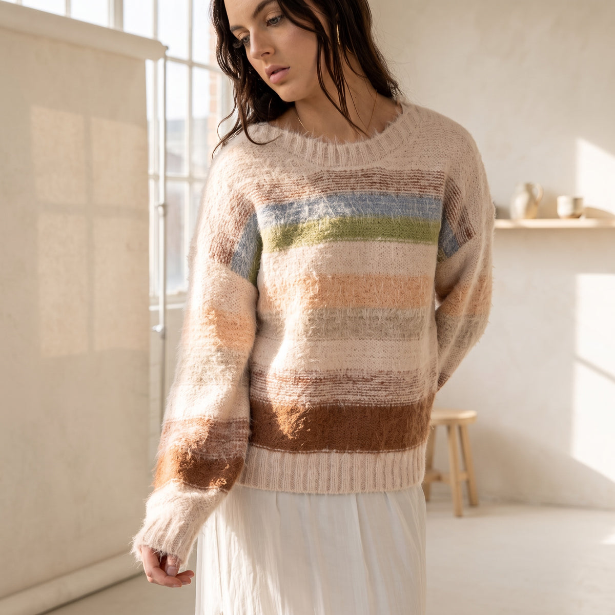 Model wearing an OKAKIE plush pastel striped sweater with a crew neckline and drop shoulders in a sunlit studio.