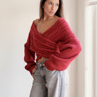Model wearing an OKAKIE red cable knit sweater with a criss-cross V-neckline and wide dolman sleeves.