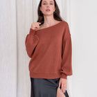 Model wearing an OKAKIE rust-colored boat-neck knit sweater with voluminous sleeves and a relaxed, draped fit.
