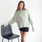 Model wearing an OKAKIE sage green fuzzy mock-neck tunic with drop shoulders and long sleeves against a white background.