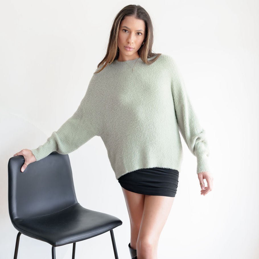 Model wearing an OKAKIE sage green fuzzy mock-neck tunic with drop shoulders and long sleeves against a white background.