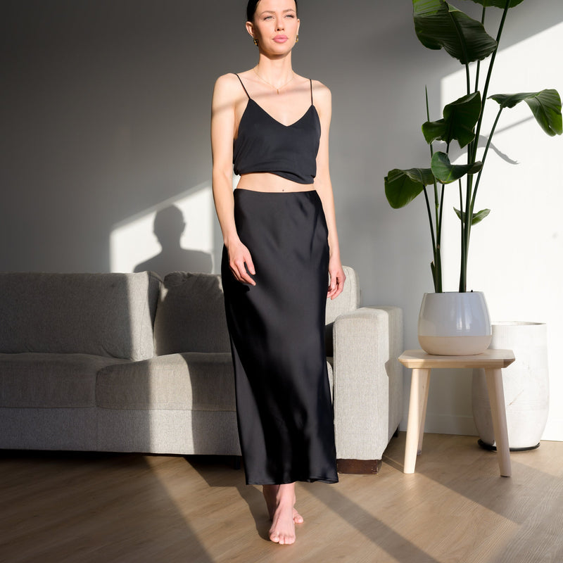 OKAKIE Serène matte satin black maxi skirt worn by a model in a sunlit room with a sofa and indoor plant.
