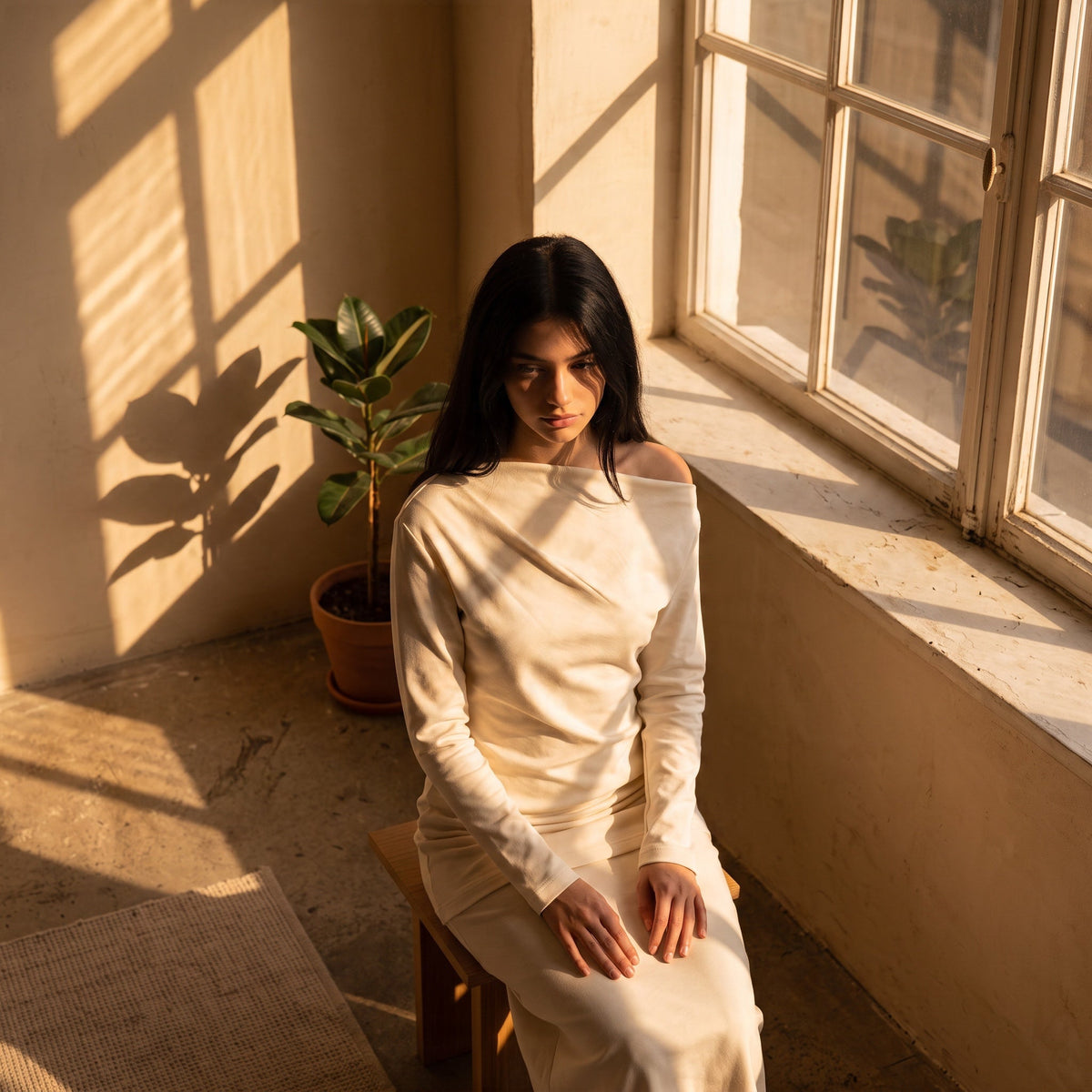 OKAKIE Solène asymmetric scuba knit top in cream worn by a model sitting by a sunlit window.