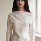 OKAKIE Solène asymmetric off-shoulder top in cream cotton-rich scuba knit with long sleeves and architectural neckline