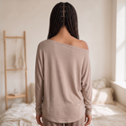 Model wearing OKAKIE beige hacci knit top with wide boat neckline and dolman sleeves, back view in bedroom setting.
