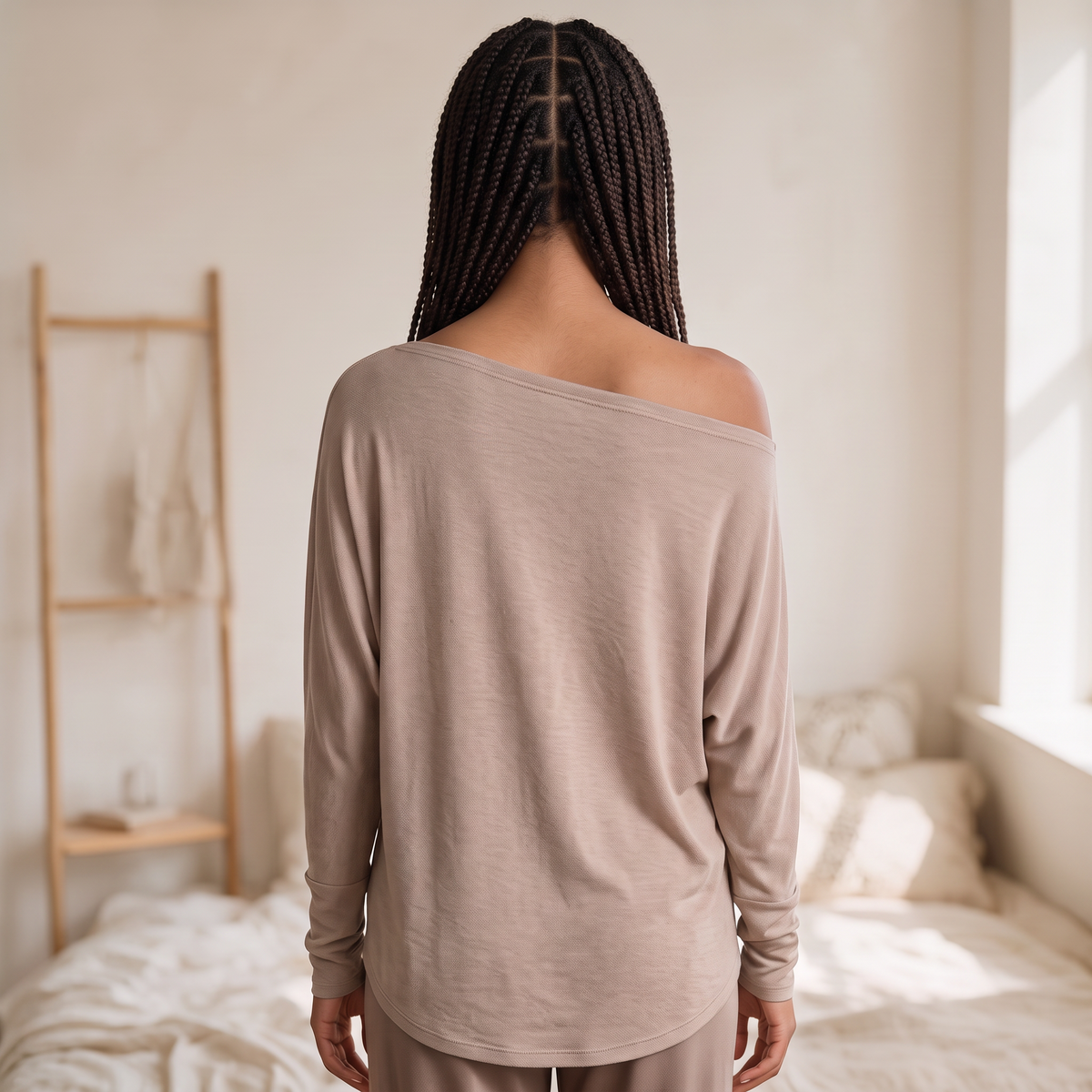 Model wearing OKAKIE beige hacci knit top with wide boat neckline and dolman sleeves, back view in bedroom setting.