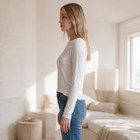OKAKIE white long-sleeve boat neck top in soft brushed knit worn by a woman in a bright, minimalist bedroom.