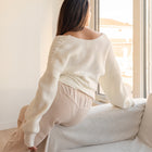 OKAKIE white cable knit sweater with dolman sleeves and off-shoulder design worn by a woman sitting on a sofa