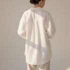 Back view of a model wearing an OKAKIE white cotton poplin boyfriend over-shirt with a loose fit and high-low hem.