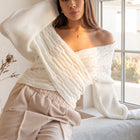 OKAKIE white cable knit sweater with a criss-cross V-neckline and dolman sleeves worn off-shoulder by a model.
